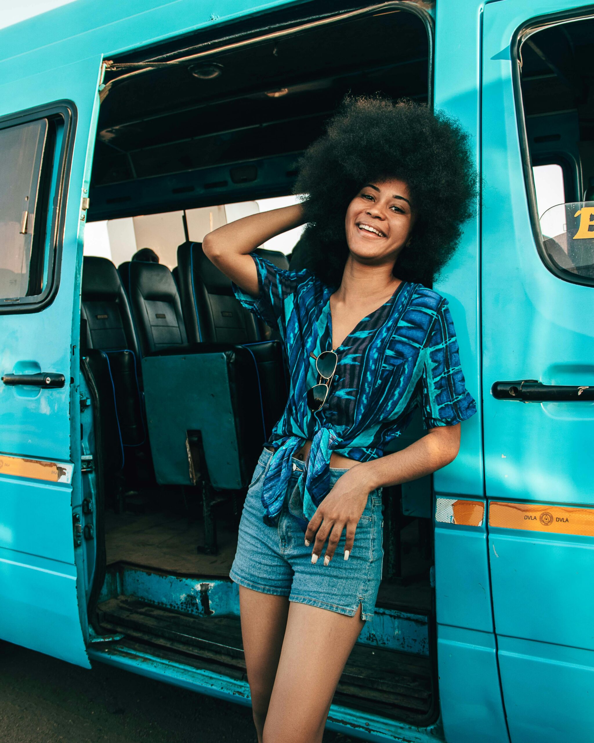 Cheerful woman with afro hair poses by a blue van, expressing joy and style.