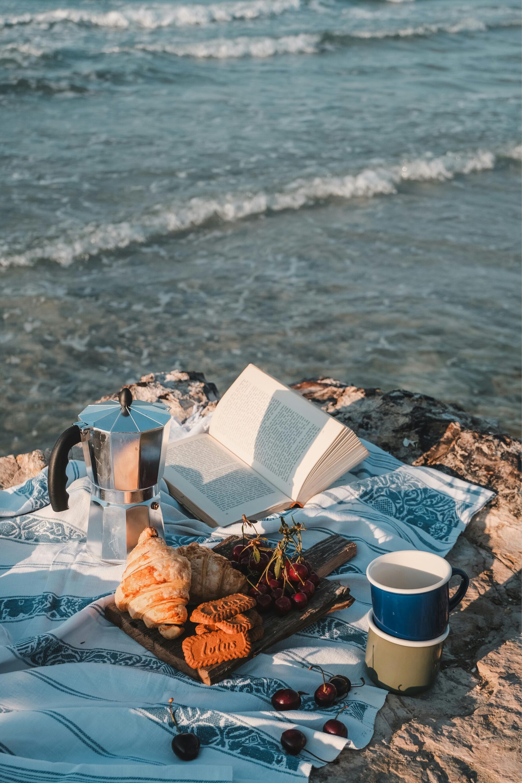 Relaxing beach picnic setup with coffee, snacks, and an open book by the sea.