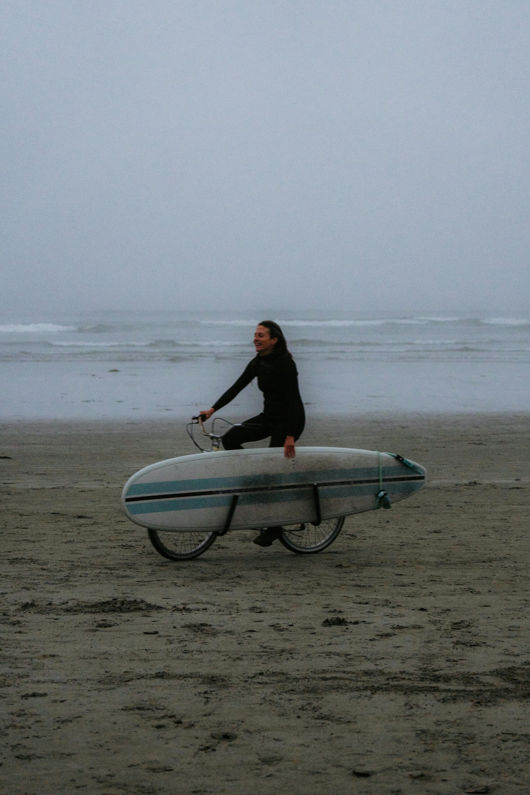 Woman cycling along a foggy beach with a surfboard, embracing adventure and freedom.
